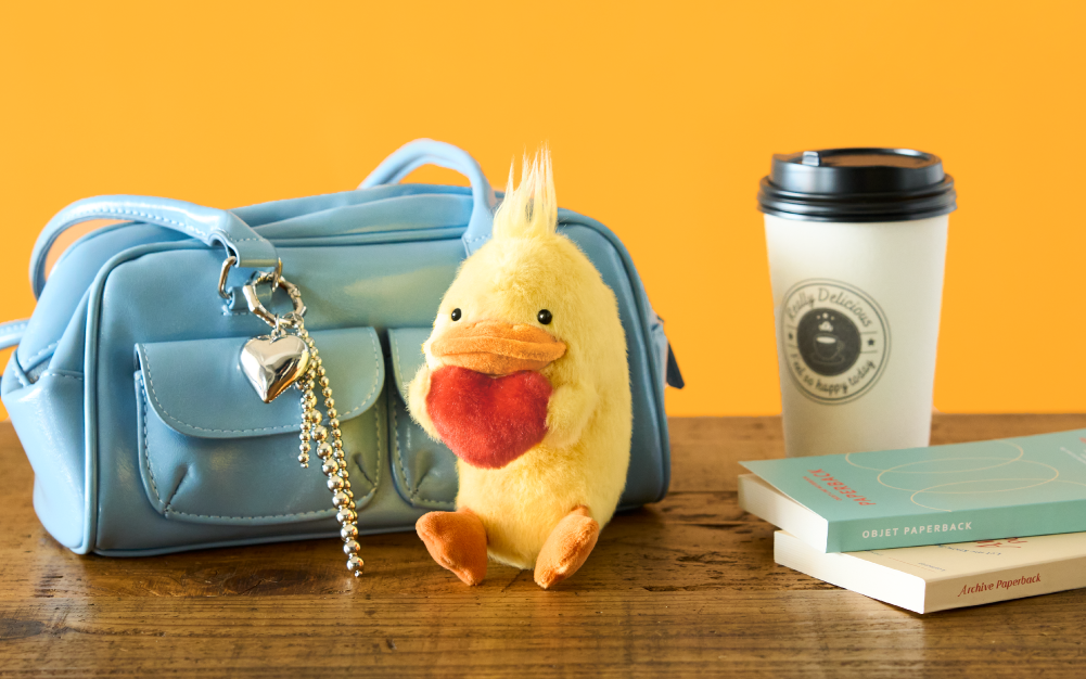 Set on a bright yellow background, atop a wooden table sits a blue leather bad, the Duck Posture Pal Bag Charm holding its red plush heart sits beside two paperback books and a to-go coffee
