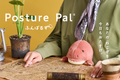 White text in English and Japanese "Posture Pal" across image. Image set with yellow background. Figure in green button down shirt with patterned tie sits at a desk looking at a vintage map with a telescope. Light red and white T-Rex Posture Pal sits in the figure's lap. Potted plant, lantern and mug sit around the figure on the desk.