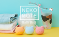 nekomote bath ball banner image featuring three nekomote bath balls (orange, blue, pink) removed from their packaging and displayed on a yellow surface in front of three towels, a loofah, and a cup shaped like a cat paw with a toothbrush inside. the product logo with the words NEKOMOTE BATH BALL and untranslated japanese text are overlaid across the image