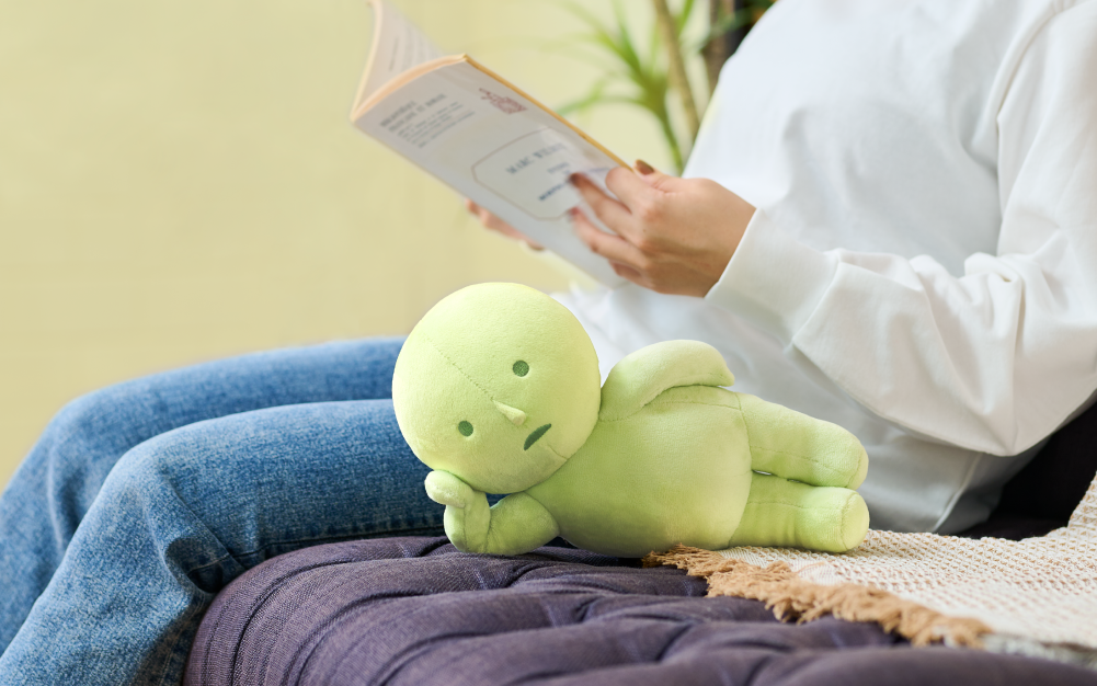 "Laying Down" SMISKI plush lays on its side, with head propped up with hand beside figure in jeans and a white shirt reading a book. Both the figure and SMISKI plush are atop a dark blue couch.