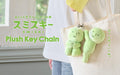 Image shows the two variations of product SMISKI Plush Key Chain written in both English and Japanese. (Right) depicts both variations of SMISKI clipped to a white canvas bag. SMISKI Hugging Knees to the left and SMISKI Hanging On to the right.